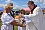 Na pierwszym planie po lewej stronie widać kobietę w białej, ludowej sukience z haftowanymi kwiatami w wianku z kwiatów i ziół. Trzyma ona wiklinowy koszyk, bogato ozdobiony wstążkami i koronkami, zawierający różne produkty, który przekazuje księdzu. Za nią stoi mężczyzna niosący bochen chleba. W tle widać inne osoby, namioty i stragany.