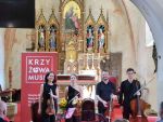 Zdjęcie przedstawia kwartet muzyków klasycznych wewnątrz zabytkowego, sakralnego wnętrza kościoła w Krzyżowej. Na pierwszym planie, od lewej, stoją cztery osoby. Trzy z nich grają na instrumentach smyczkowych - dwie skrzypaczki i wiolonczelista. Czwarta osoba, mężczyzna z brodą, trzyma altówkę. Wszyscy ubrani są na czarno. W tle, za muzykami, widoczny jest bogato zdobiony, złoty ołtarz z figurami świętych. Na środku ołtarza znajduje się obraz przedstawiający Matkę Boską z Dzieciątkiem. Po lewej stronie, obok muzyków, stoi duży, czerwony baner z napisem Krzyżowa Music.